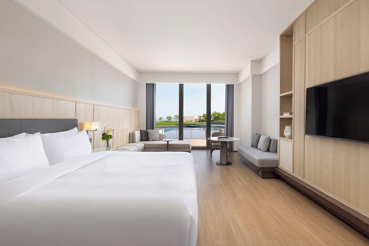 A hotel room with a large bed, a TV, a sofa, a small table, and a view of a pool.