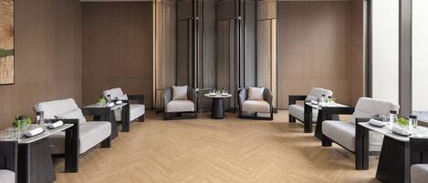A hotel room with a herringbone floor, a large window, a seating area with tables and chairs, and a decorative wall panel.