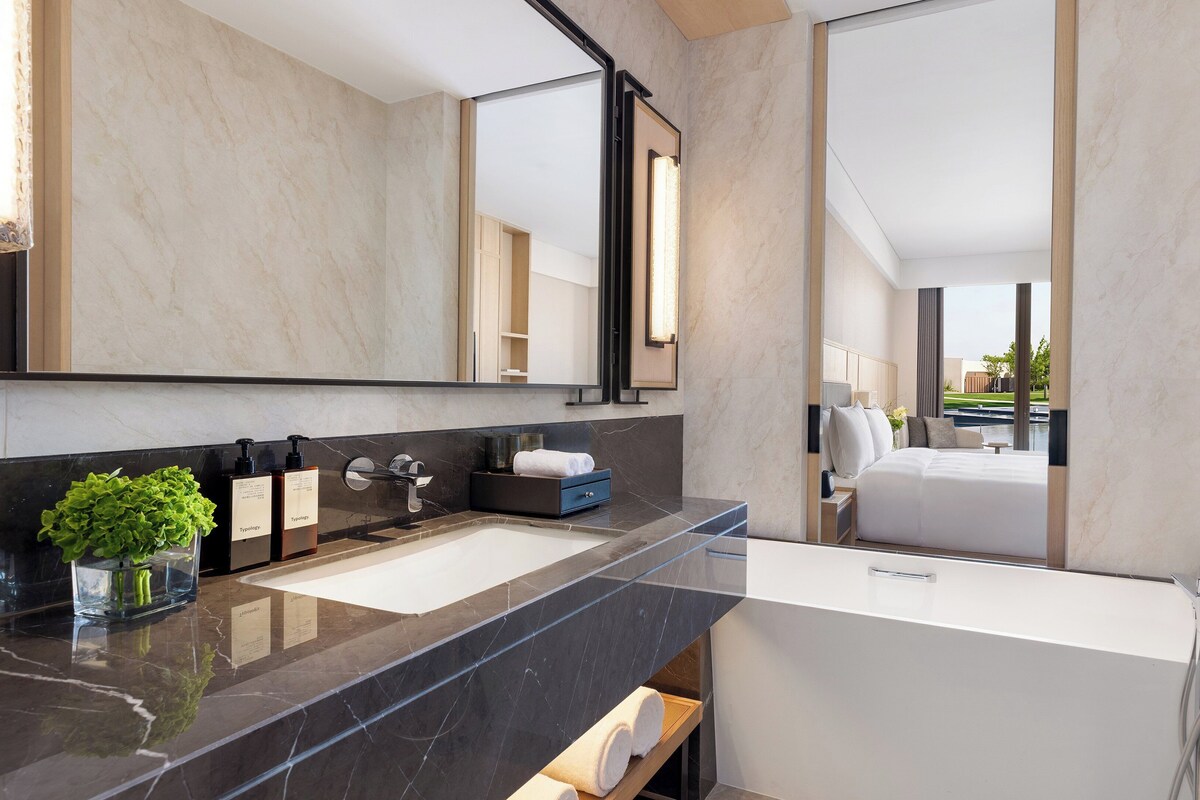 Une salle de bain moderne dotée d’un double lavabo, d’un grand miroir et offrant une vue sur une chambre avec un lit et une piscine.