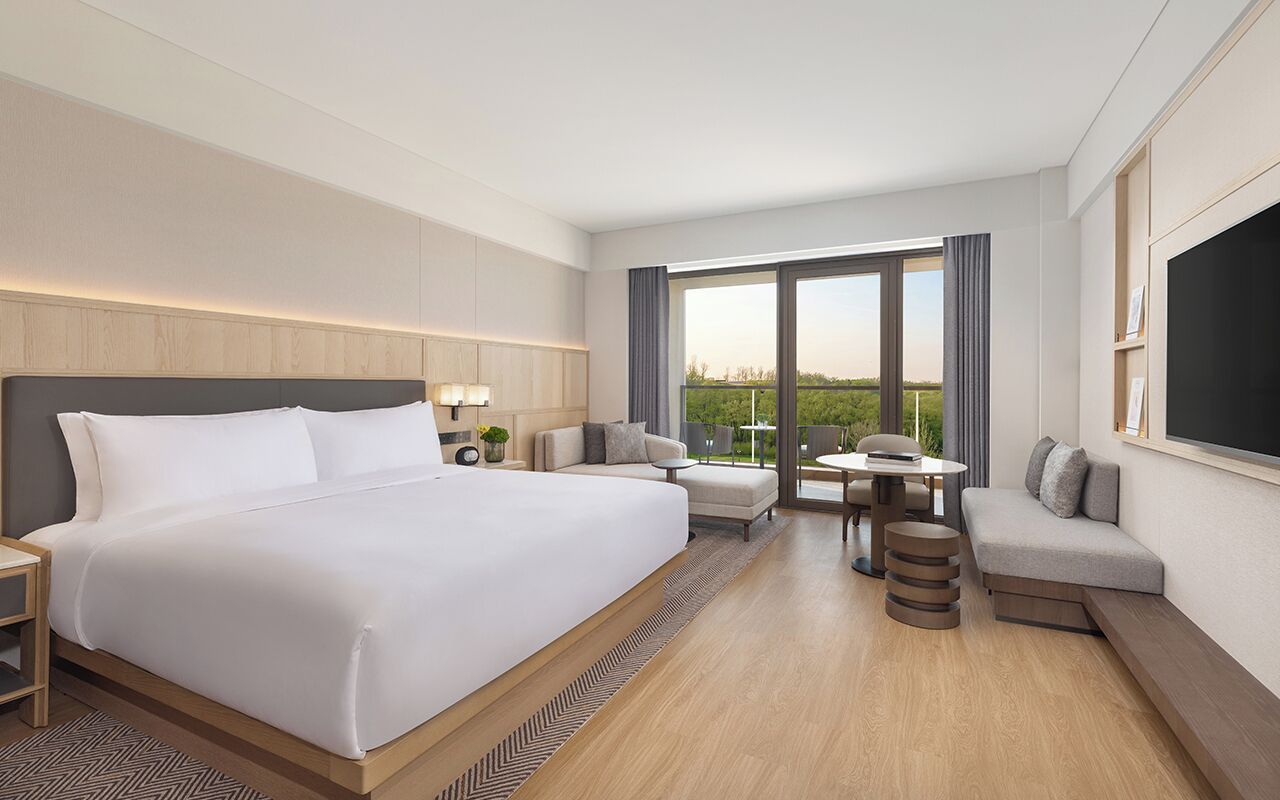 A hotel room with a large bed, a seating area with a table, and a view of greenery outside.
