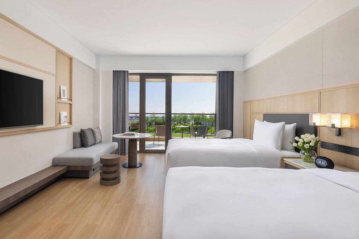 A hotel room with two beds, a TV, a small table, and a balcony with a view.