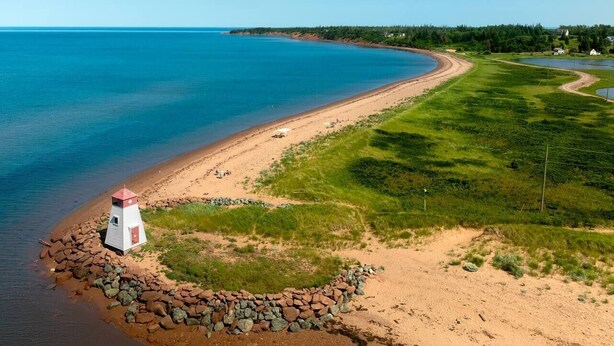 A coastal landscape with a lighthouse, sandy beach, grassy areas, and a clear blue sky.