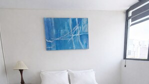 Ein ordentlich bezogenes Bett mit weißen Leinen in einem Zimmer, das durch ein blaues, abstraktes Gemälde an der Wand und ein Fenster rechts gekennzeichnet ist.