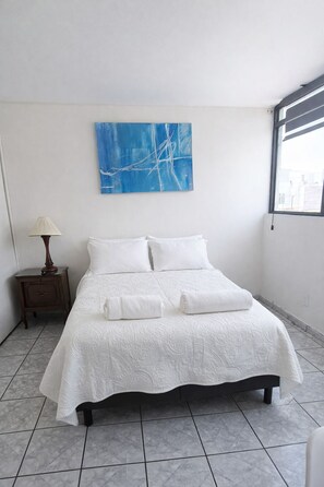 Una cama bien hecha con sábanas blancas en una habitación con un cuadro abstracto azul en la pared y una ventana a la derecha.