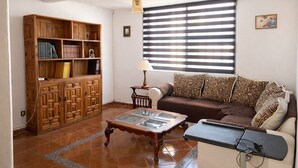 Ein Wohnzimmer mit einer Holzbücherreolen, einem Glastisch, einem braunen Sofa, einem gemusterten Kissen, einer Lampe und einem Fenster mit Jalousien.