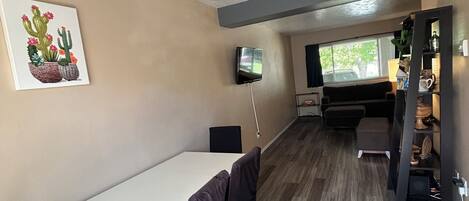 Une pièce dotée d’un ventilateur de plafond, d’une télévision, d’une table à manger avec des chaises, d’une fenêtre avec des stores et d’un tableau représentant un cactus accroché au mur.