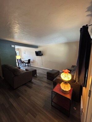 A living room with a sofa, ottoman, lamp, flat-screen TV, and a dining area in the background. - Cute 2 bedroom condo, centrally located in Boise (Boise)