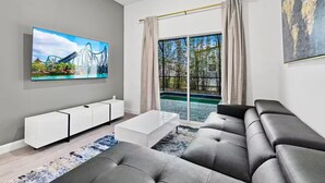A modern living room with a large flat-screen TV, a grey sofa, a white coffee table, and a large window with a view of a pool and trees. - The Epic Retreat by Disney (Davenport)