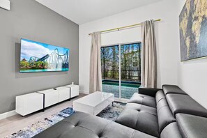Uma sala de estar moderna com uma grande televisão de tela plana, um sofá cinza, uma mesa de centro branca e uma janela ampla com vista para uma piscina e árvores.