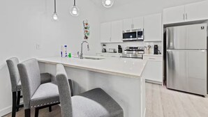 A modern kitchen with white cabinetry, a stainless steel refrigerator, a microwave, and a sink. There are two bar stools and pendant lights above the counter. - The Epic Retreat by Disney (Davenport)