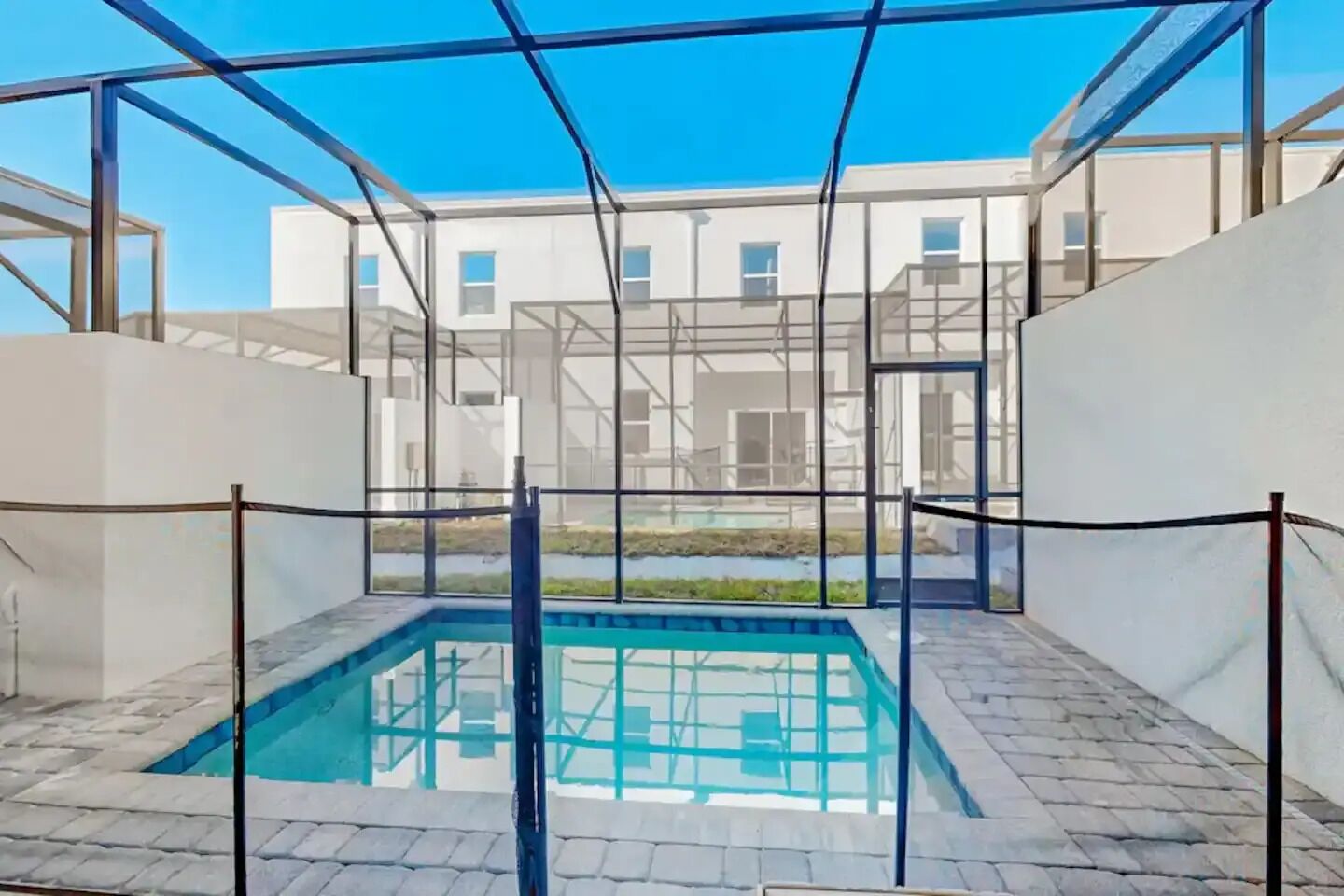 A swimming pool with a glass enclosure, surrounded by a paved area and a building in the background.
