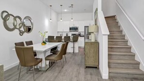 A modern kitchen with white cabinets, a dining area with a white table and brown chairs, and a wooden staircase. - Serendipity @Azur Retreat by Disney w private pool (Davenport)