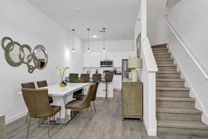 Eine moderne Küche mit weißen Schränken, ein Essbereich mit weißem Tisch und braunen Stühlen sowie eine Holztreppe.