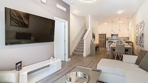 Un soggiorno moderno con una televisione a schermo piatto, un divano, un tavolino da caffè e una scala che conduce a una cucina.