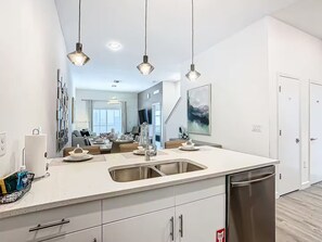 Et moderne køkken med en central ø, rustfrie stål-apparater, hængende lamper og et spiseområde med udsigt til stuen.