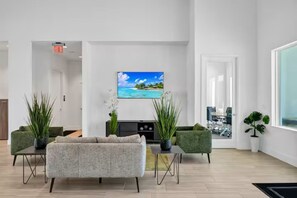 Ein modernes Wohnzimmer mit einer Couch, zwei grünen Sesseln, einem Couchtisch, einem an der Wand montierten Fernseher und Topfpflanzen.