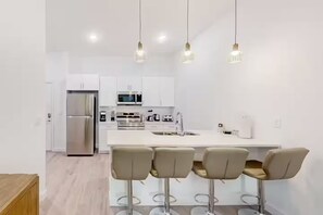 A modern kitchen with white cabinets, stainless steel appliances, a central island with bar stools, and pendant lighting. - The Onyx Retreat by Disney / Universal/ Seaworld (Davenport)