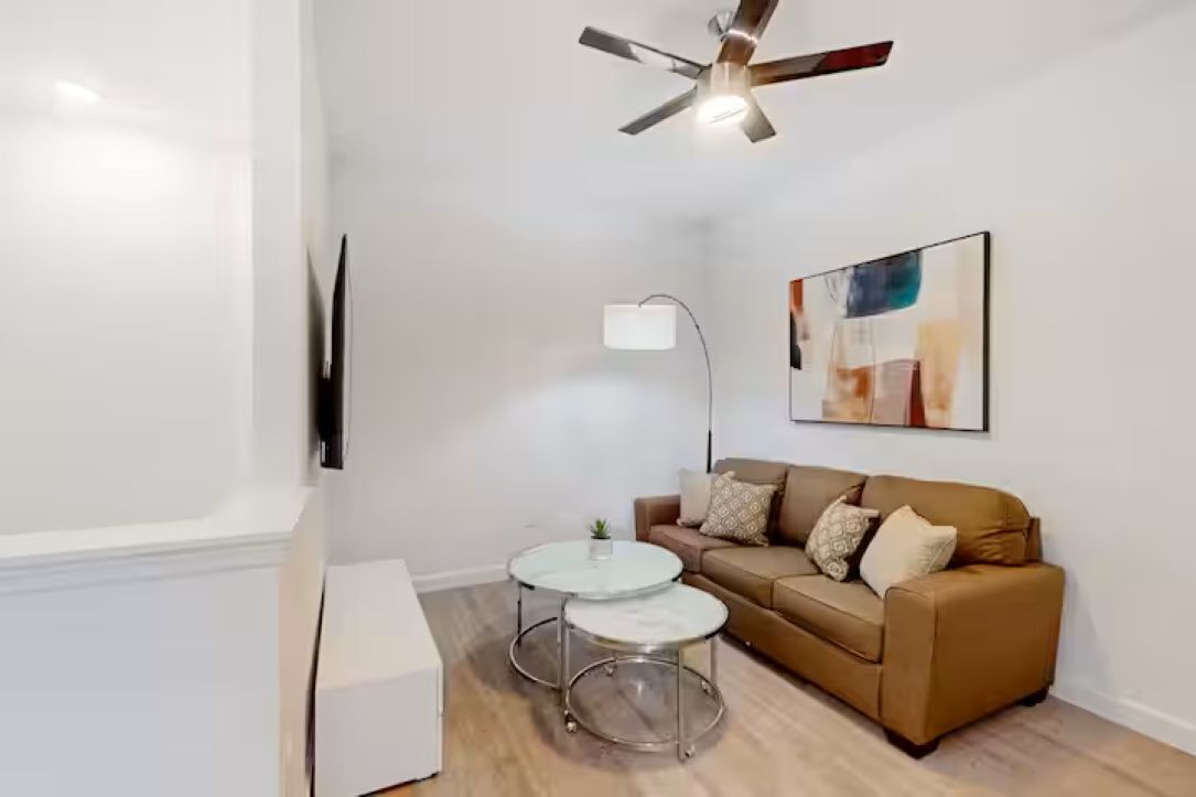 A modern living room with a brown sofa, two round tables, a ceiling fan, a mounted TV, and a painting on the wall.