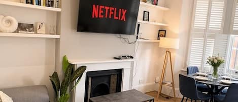 A modern living room with a grey sofa, a fireplace, a dining area with a table and chairs, and a TV mounted on the wall displaying 'NETFLIX'.