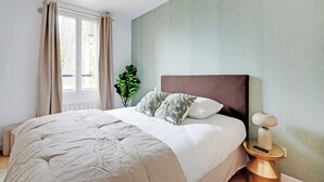 Um quarto com cama, mesa de cabeceira com abajur, janela com cortinas e um vaso de planta.
