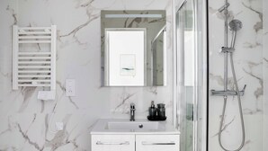 Uma casa de banho com toucador branco, espelho, chuveiro com chuveiro manual e porta-toalhas.
