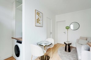 A compact living space with a washing machine, a dining table with chairs, a round coffee table, a sofa, a mirror, and a framed artwork on the wall. - Cozy Apartment 1br/3p - Ivry (Ivry-sur-Seine)