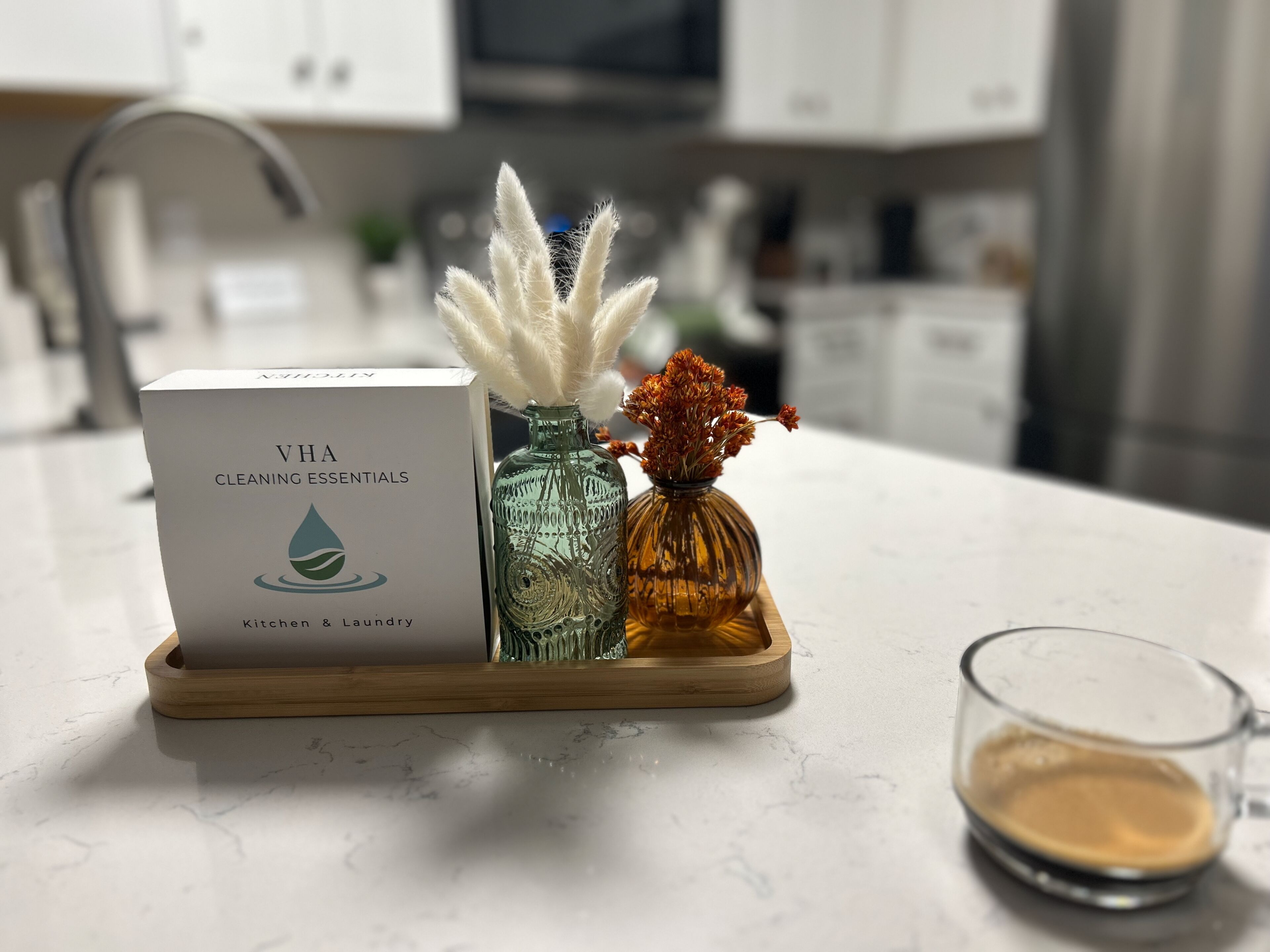 A kitchen counter with a box labeled 'VHA Cleaning Essentials,' two glass vases with dried flowers, and a glass of coffee.