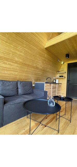 A cozy room with a wooden ceiling, a black sofa, a small round table, and a kitchen area with a sink and shelves.