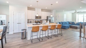 A modern kitchen with white cabinets, a central island with bar stools, a dining area with a wooden table, and a living room with a blue sofa. - Coastal Townhome with 2 Pools & Gym (Berlin)