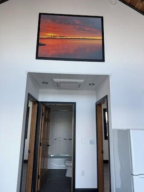 Un pasillo con un cuadro enmarcado de un atardecer, una puerta que da a un baño y un refrigerador blanco.