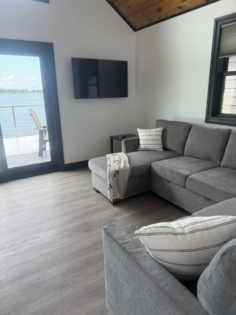 A modern living room with a grey sectional sofa, a flat-screen TV mounted on the wall, and a large window overlooking a body of water.