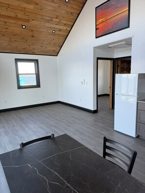 A spacious room with a wooden ceiling, a dining table, a refrigerator, and a window.