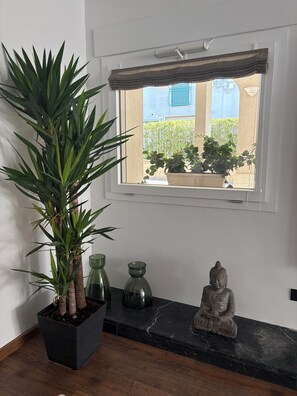 Ein Zimmer mit einer großen Topfpflanze, zwei Glasvasen, einer Buddha-Statue und einem Fenster mit Blick auf ein blaues Gebäude und Grünflächen.