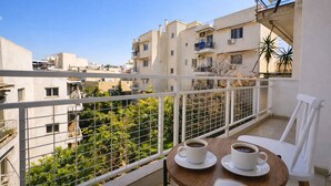 Un balcón con dos sillas blancas y una mesa, dos tazas de café y vistas a edificios y vegetación.