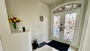 Een hal met een witte deur met decoratieve glaspanelen, een kleine vaas met bloemen, een potplant en een metalen bankje.
