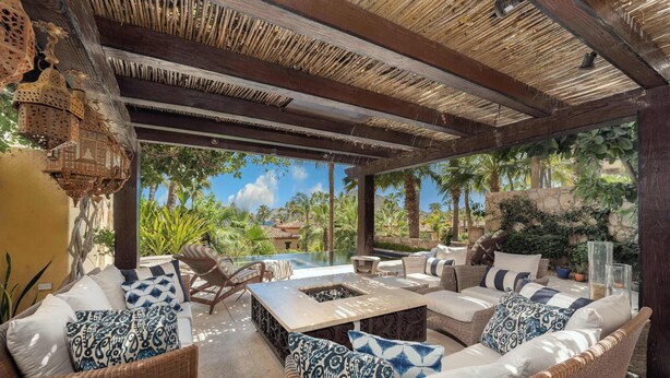 Ein Außenbereich mit Loungemöbeln aus Rattan, einer Feuerstelle und Blick auf Pool und Grünflächen.