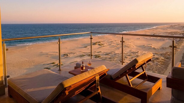 Uma varanda com vista para a praia, cadeiras de descanso e uma mesa com garrafa e copo.