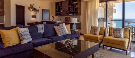 A modern living room with a sofa, armchairs, a glass coffee table, and a kitchen area with wooden cabinets.