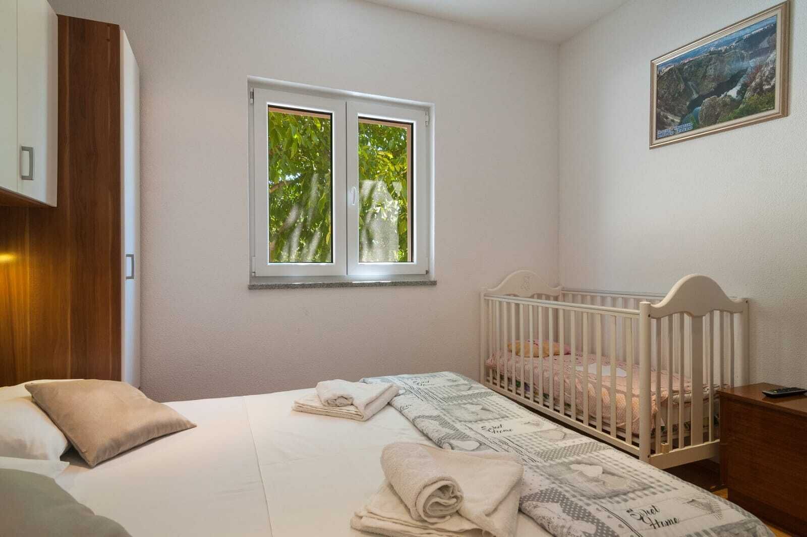 A bedroom with a crib, a bed with pillows, a window with a view of trees, and a painting on the wall.