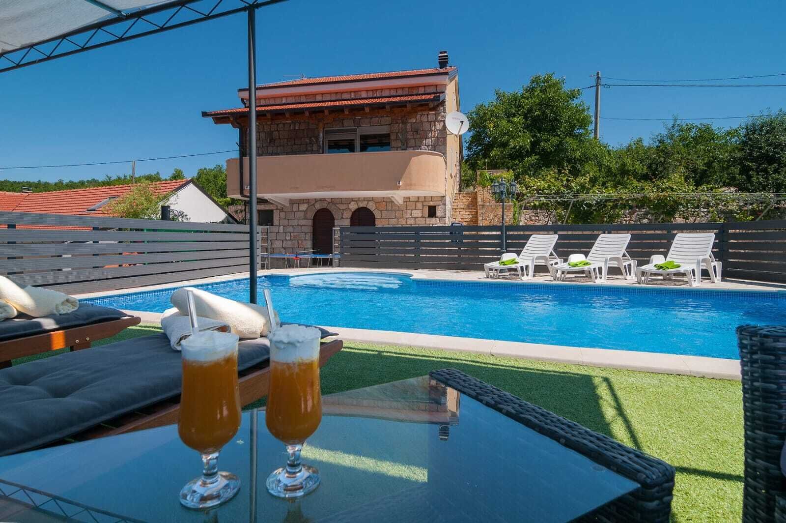 Un área de piscina con sillones de descanso, una mesa con bebidas y un edificio de piedra al fondo.
