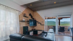 En moderne stue med en sort lædersofa, et fladskærms-tv, en murstensvæg og en skydedør, der fører til et udendørs område.