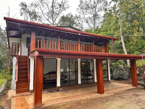 Una casa de madera de dos pisos, con balcón, ventanas grandes y una terraza de madera.