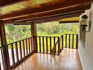 A wooden balcony with a railing, a view of greenery, and a wall-mounted light fixture.