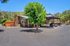 Un chalet doté d’une cheminée en pierre, d’un patio couvert et d’un arbre devant.