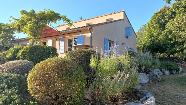 Een gebouw met een schuin dak, meerdere ramen met luiken en een verzorgde tuin met verschillende struiken en een stenen pad.