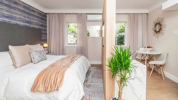 Ein Schlafzimmer mit Bett, Nachttisch, Vorhängen, einem Fenster, einer Pflanze und einem Essbereich mit Tisch und Stühlen.