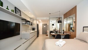Et moderne hotelværelse med et fladskærms-tv, en seng med pænt foldede håndklæder, et spiseområde med bord og stole samt et tekøkken med mikroovn og vask.