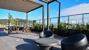 An outdoor seating area with modern chairs and a bench, surrounded by potted plants and a view of a cityscape. - Studio em frente a UFSC e HU - 309A (Florianópolis)