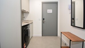 A laundry area with a washing machine, a grey door with a sign, and a wooden bench. - Studio em frente a UFSC e HU - 309A (Florianópolis)