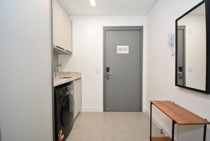 A laundry area with a washing machine, a grey door with a sign, and a wooden bench. - Studio em frente a UFSC e HU - 309A (Florianópolis)